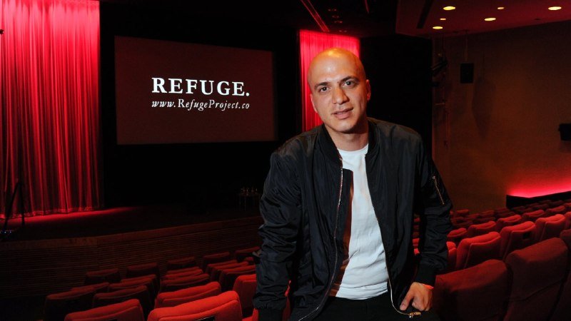 A bald man in a black bomber jacket and white t-shirt stands inside a red-lit theater, with a screen behind him displaying "REFUGE. www.RefugeProject.co."