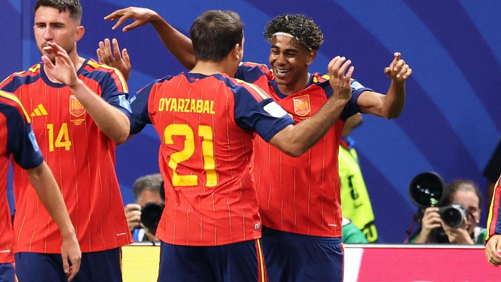 Soccer players in red and yellow Spain jerseys, including one labeled "OYARZABAL 21," celebrate together on the pitch as photographers capture the moment.