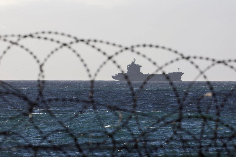 A naval warship marked "102" sails on choppy ocean waters, viewed through coils of barbed wire in the foreground.