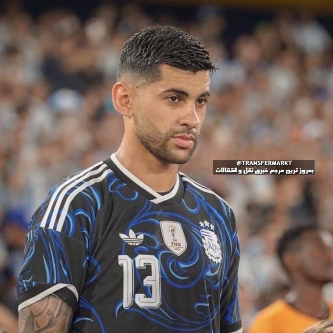 A bearded soccer player wearing a black and blue patterned number 13 jersey stands in a stadium with a blurred crowd behind him.