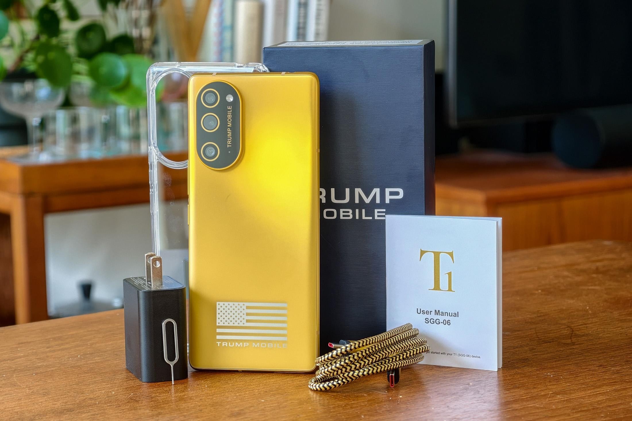 A gold Trump Mobile smartphone with an American flag logo sits on a wooden table alongside its black packaging box, charging adapter, clear case, braided cable, and T1 user manual.