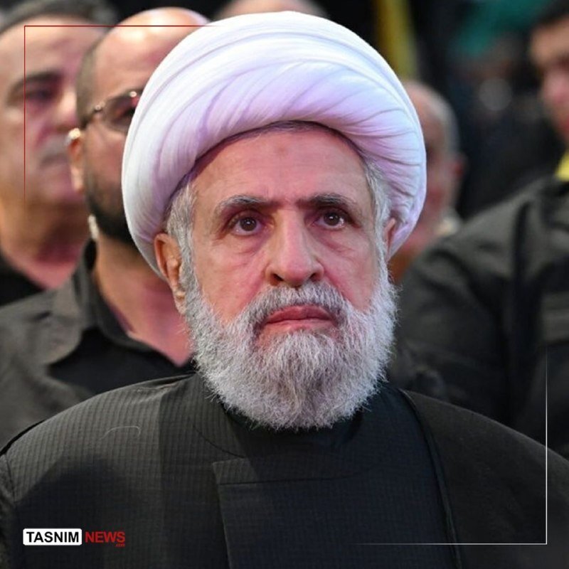 Bearded cleric in white turban and black robe stands solemnly among other men in dark clothing, with a "TASNIM NEWS" logo visible at bottom left.