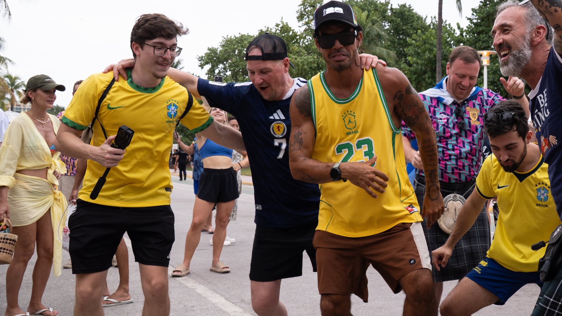 Fans in Brazil and Scotland soccer jerseys walk together arm-in-arm down a street, smiling and celebrating together.