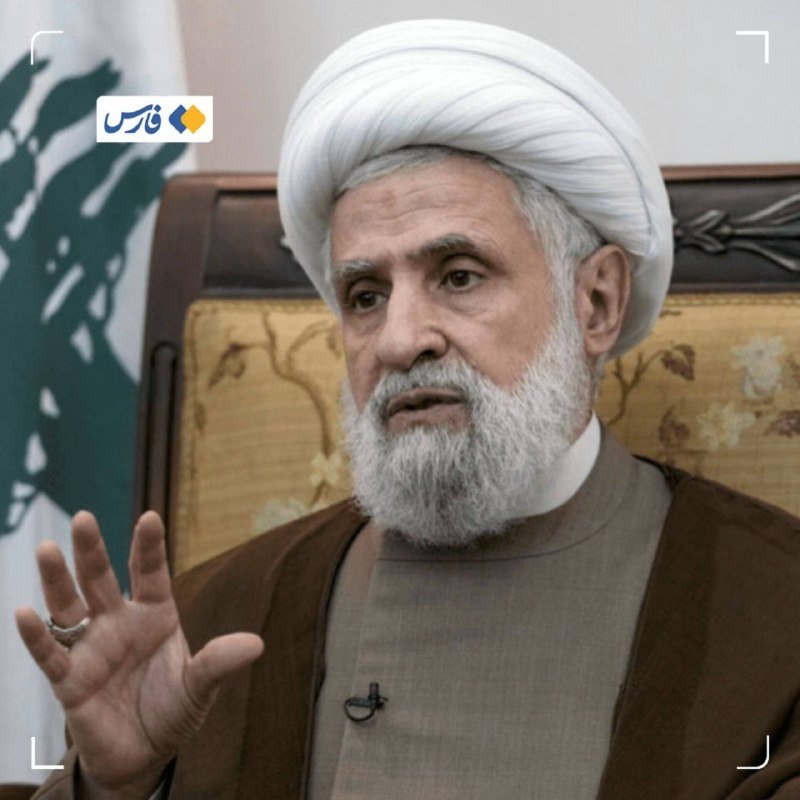 A bearded man in a white turban and brown robe speaks while gesturing with his hand, seated before a Lebanese flag.