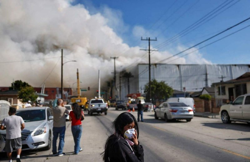 A young girl covers her face with a cloth as thick smoke billows from a large warehouse fire, while bystanders watch from a residential street lined with cars and utility poles.