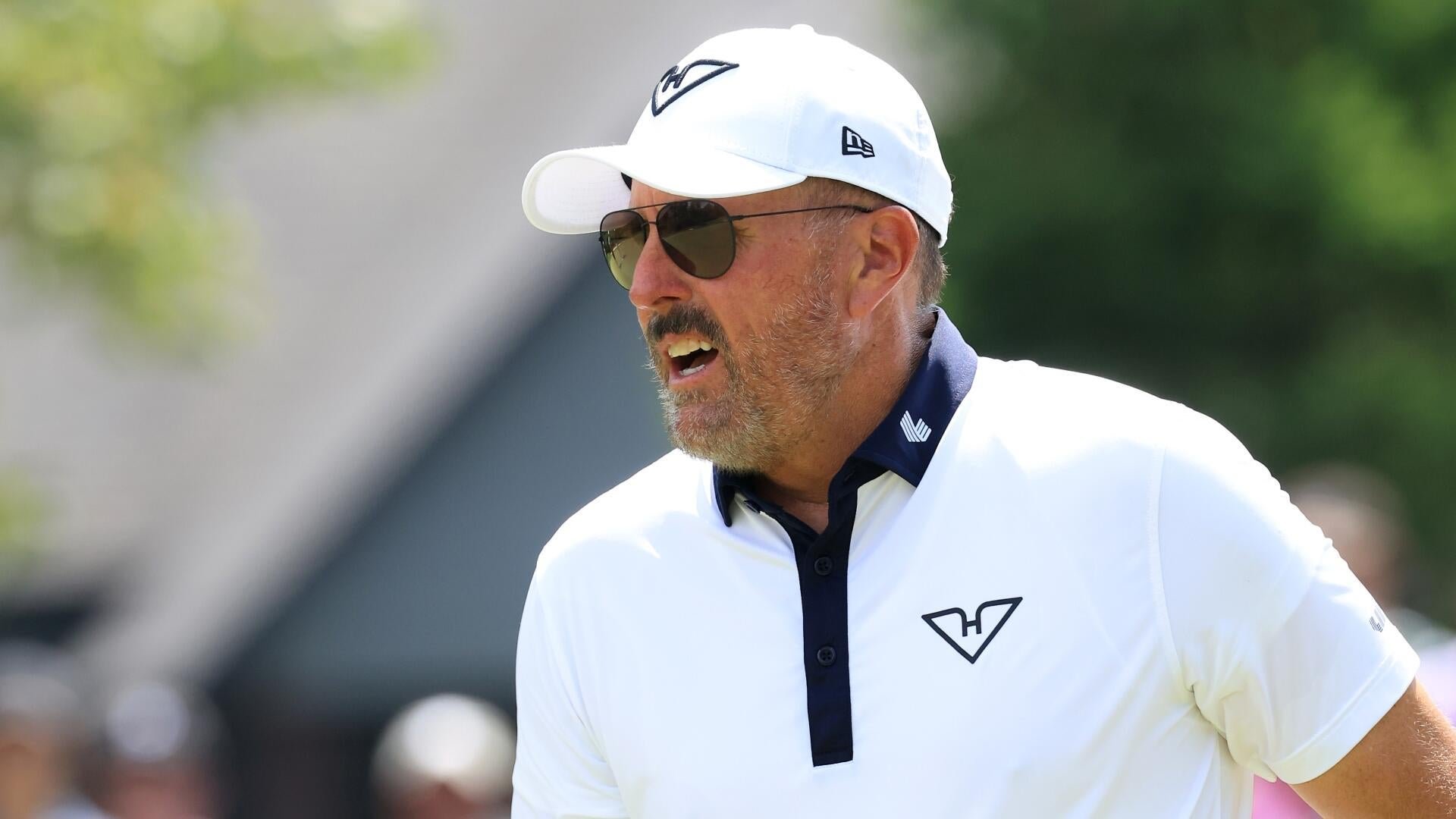 A bearded man wearing sunglasses and a white cap and polo shirt speaks outdoors, his mouth open mid-sentence against a blurred green background.