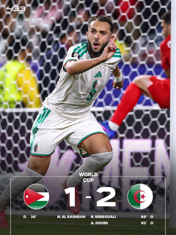 A bearded soccer player in a white and green jersey celebrates with a raised index finger in front of a goal net, while a graphic displays a World Cup match score of 1-2.