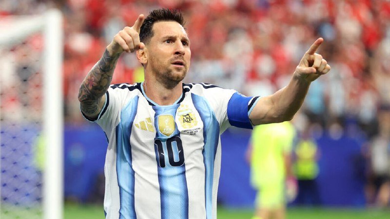 A soccer player in a light blue and white striped Argentina jersey with the number 10 points both index fingers upward, with a goalpost and crowd visible behind him.