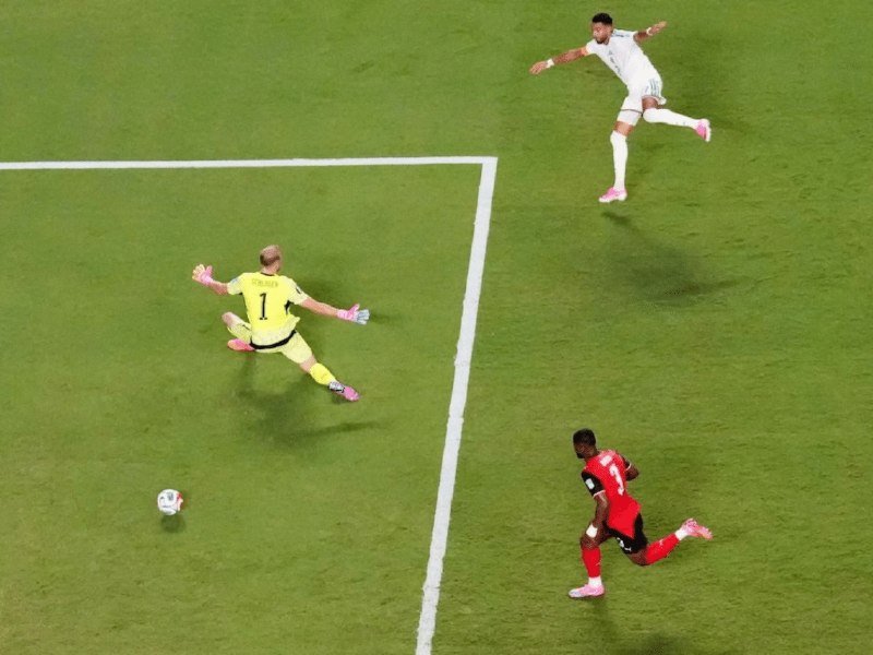 A goalkeeper in yellow dives toward the ball near the goal line as a player in red and another in white watch during a soccer match on a grass field.