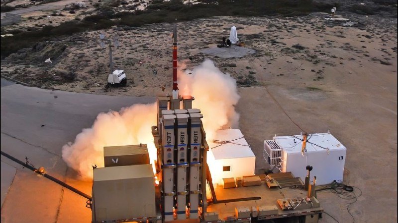 A missile launches vertically from a coastal facility, producing flames and smoke, with additional canisters and equipment visible on the launch platform.