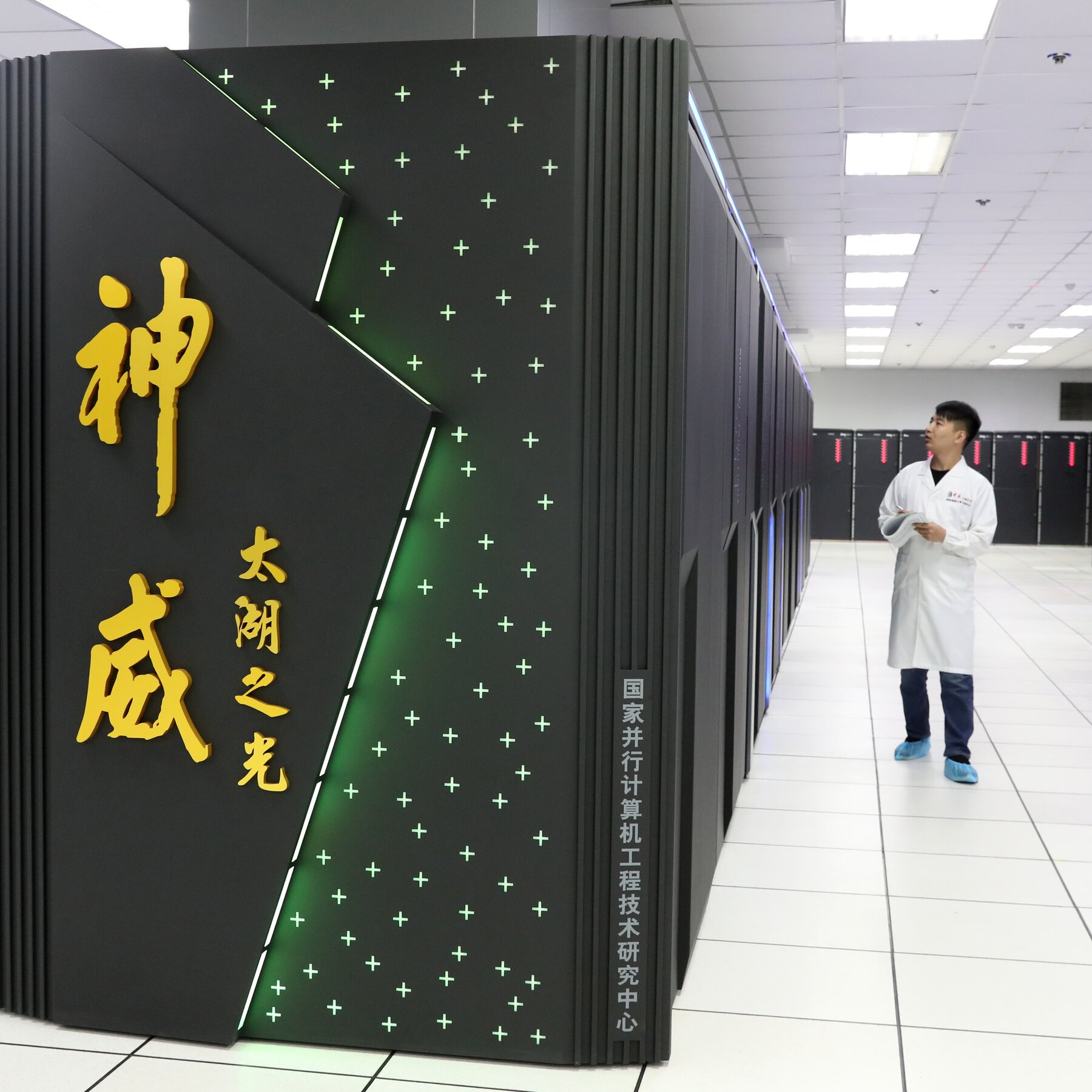 A man in a white lab coat stands beside a large black supercomputer with green illuminated plus signs and Chinese characters, inside a bright data center facility.