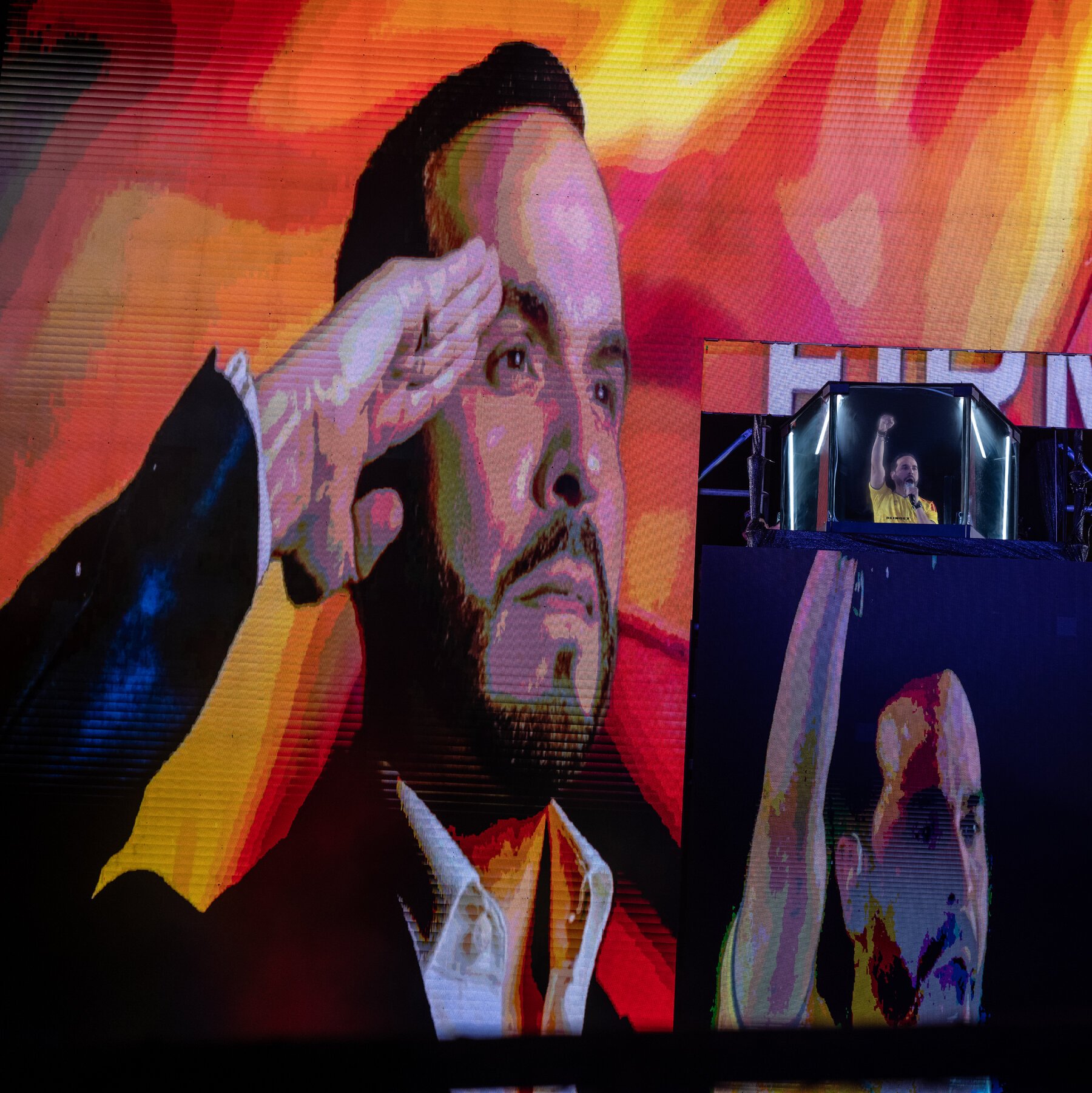 A large screen displays a portrait of a man with his hand to his head against a vivid orange and red background, while a smaller screen shows him in a yellow shirt raising his fist.