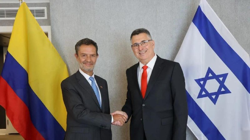 Two men in dark suits shake hands, flanked by the Colombian flag on the left and the Israeli flag on the right, against a gray curtain backdrop.