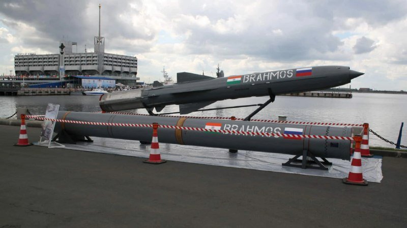 Two gray missiles labeled "BRAHMOS" with Indian and Russian flags are displayed on stands at a waterfront, cordoned by traffic cones and barriers.