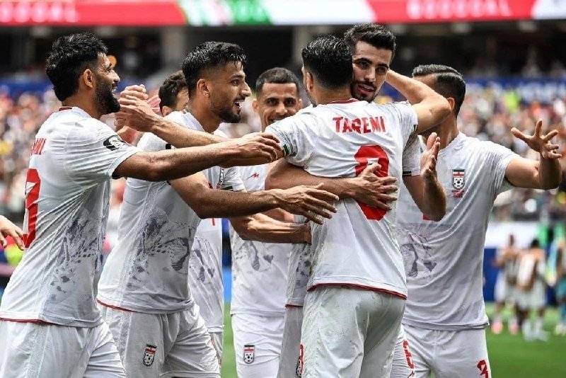 Soccer players in white jerseys with red trim embrace and celebrate together on a stadium field, with "TARE MI" and the number 9 visible on one player's back.