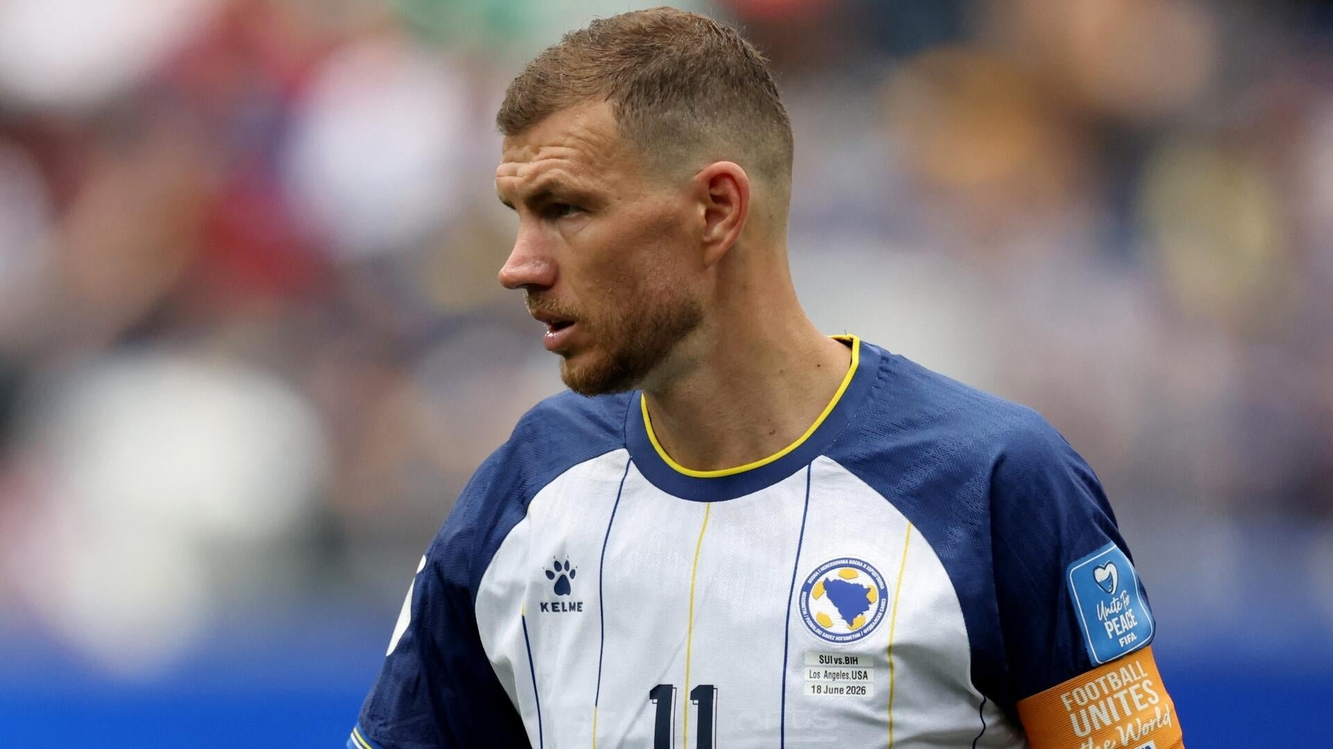 A soccer player wearing a white and navy Bosnia and Herzegovina jersey with the number 11 stands on the field, with a blurred crowd visible in the background.