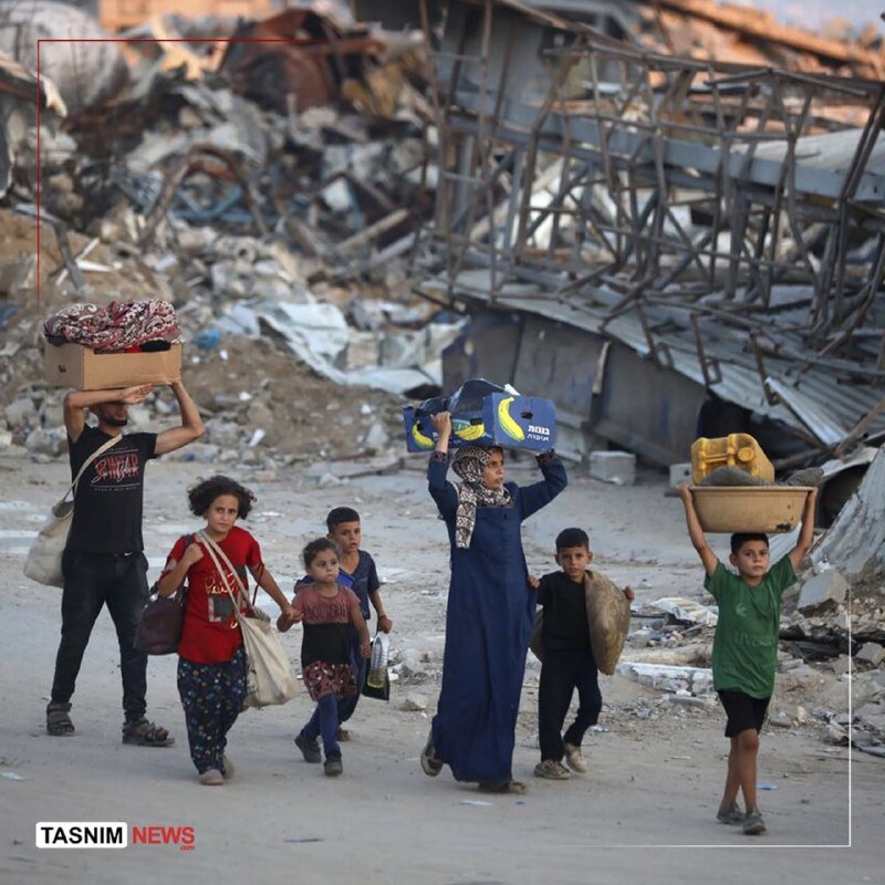 A woman in a blue dress and headscarf carries belongings on her head while walking with children along a debris-strewn street beside collapsed metal structures.