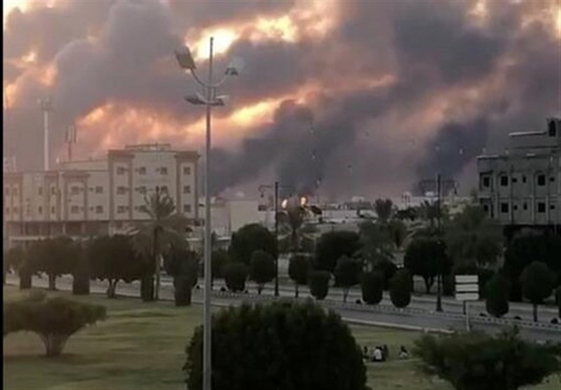 Dark smoke billows behind a row of low-rise buildings and palm trees at dusk, with a streetlamp in the foreground.
