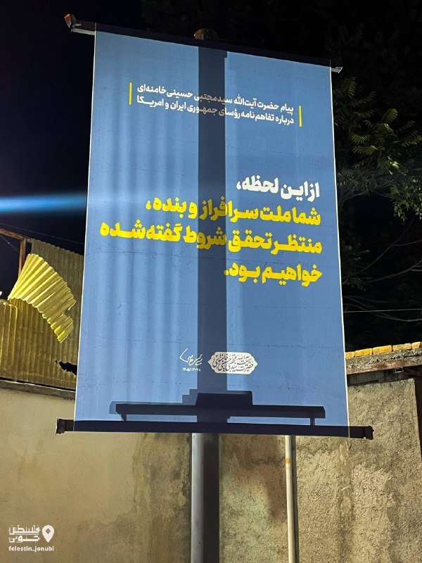 An outdoor billboard displays a blue poster with Persian text promoting an Islamic-Iranian civilization festival, mounted on a pole at night near a wall and foliage.