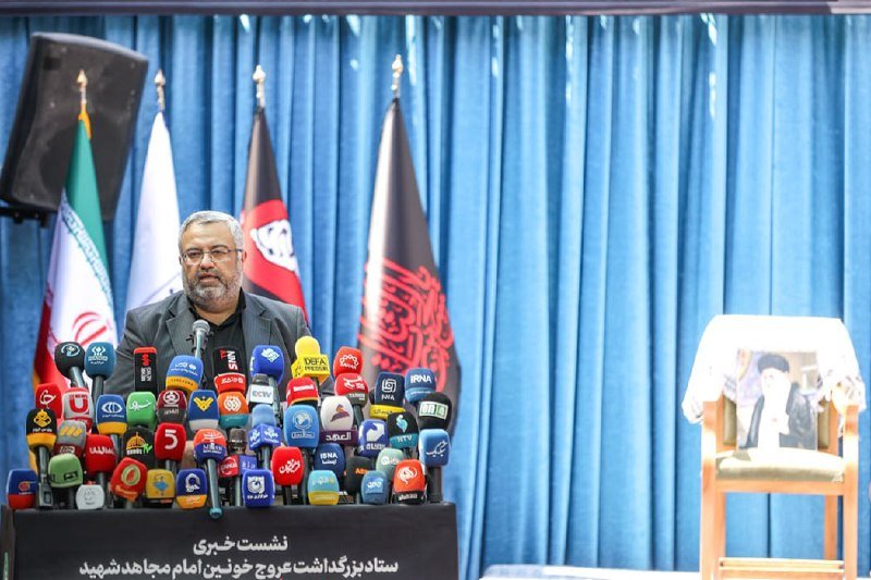 A bearded man speaks at a podium crowded with microphones from numerous media outlets, with Iranian and Afghan flags behind him and an empty chair holding a framed photo to his right.