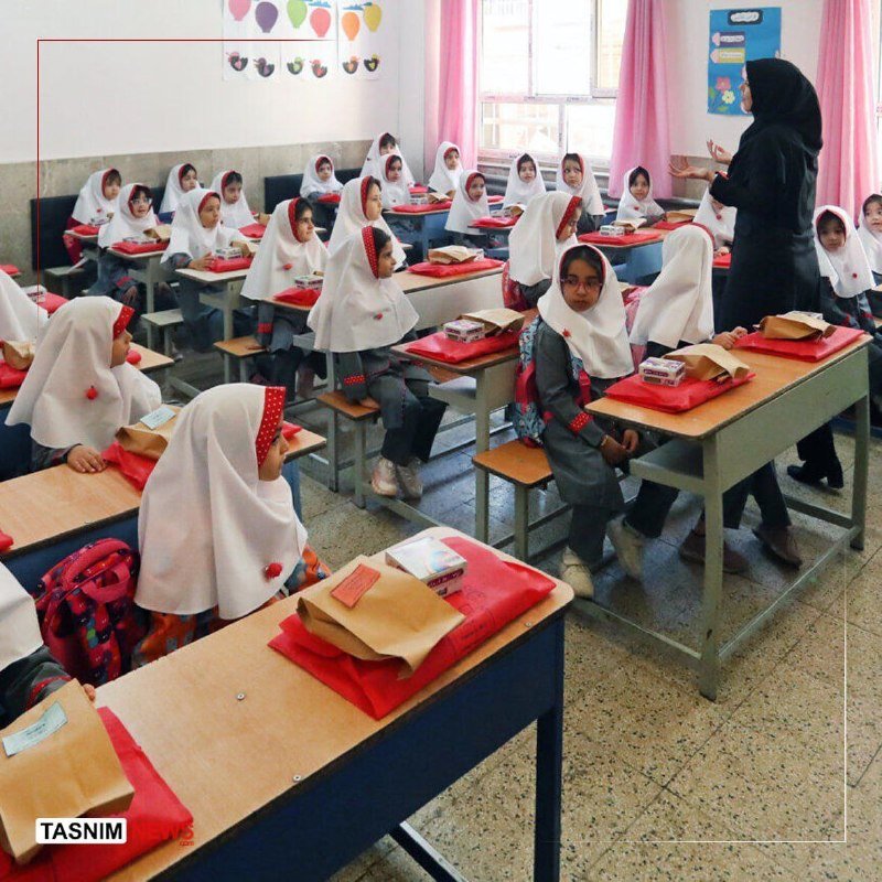 Female students in white headscarves sit at desks with red supplies while a teacher in black speaks to the class in a classroom.