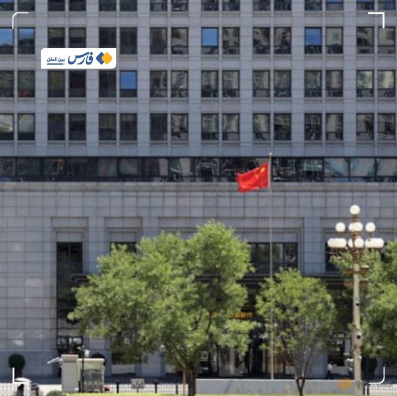 A red flag with yellow stars flies in front of a large glass-windowed office building, with green trees and a decorative streetlamp visible in the foreground.