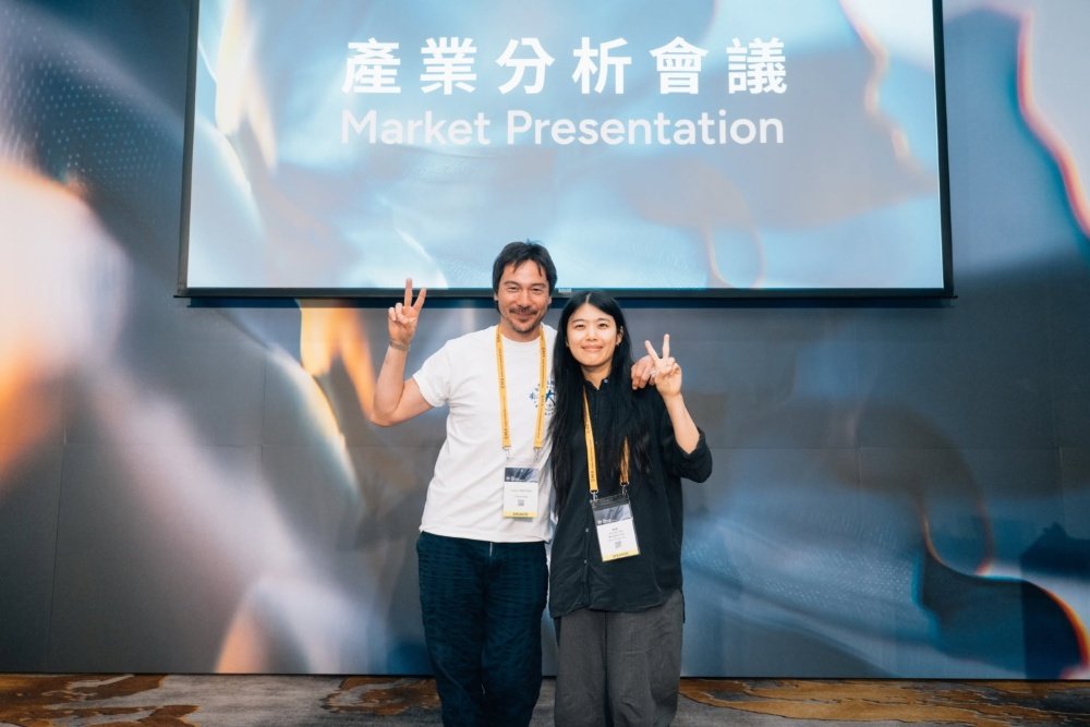 A man and woman wearing lanyard badges pose with peace signs beneath a screen displaying "產業分析會議 Market Presentation."