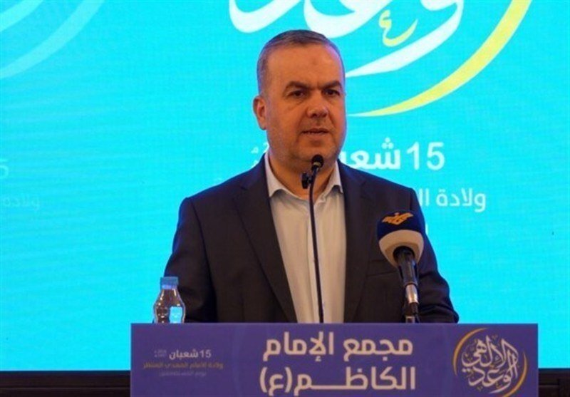 A man in a dark suit speaks at a podium with microphones, backed by a teal banner featuring Arabic text and a logo.