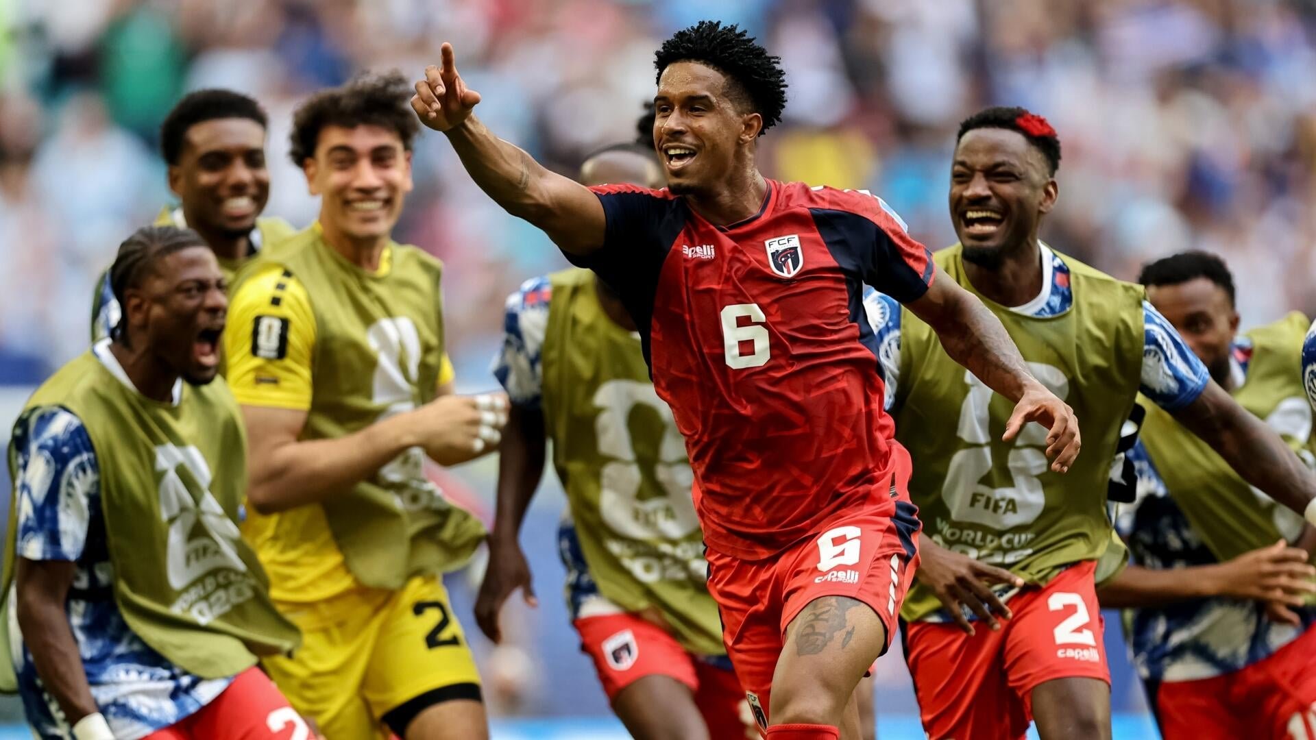 Soccer players in red and yellow training bibs celebrate on the pitch, with one player in a red jersey marked "6" pointing upward as teammates cheer around him.