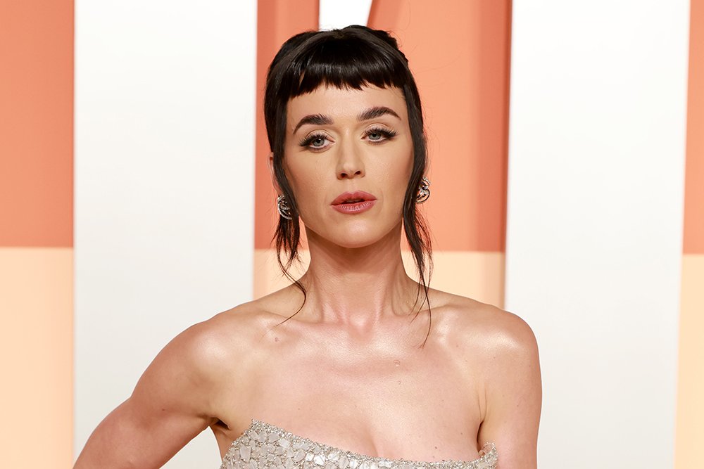 A woman with dark hair and blunt bangs poses in a strapless, jewel-encrusted gown against an orange and white background.