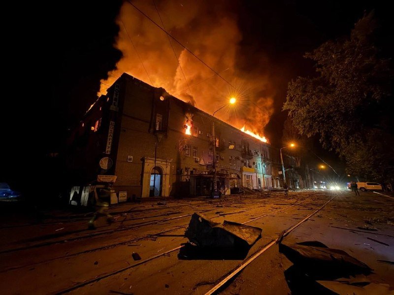 Flames engulf a multi-story building at night, with debris scattered across the street and smoke billowing into the sky.