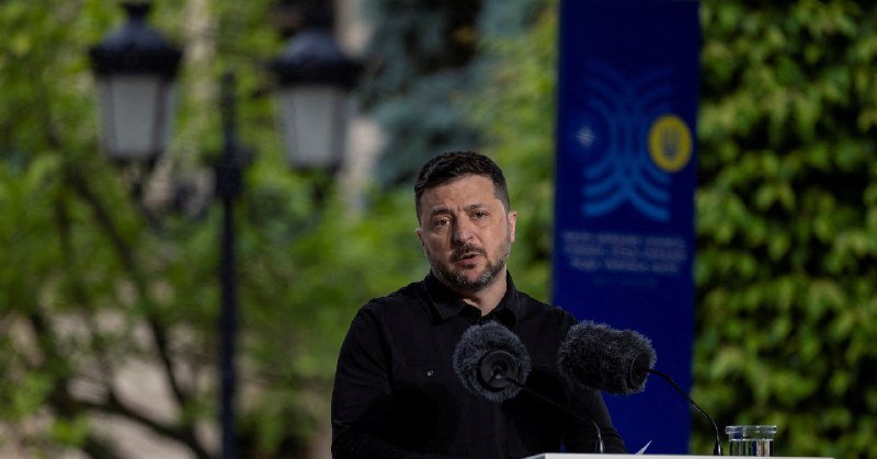 A man with short dark hair and a beard speaks at a podium with two microphones, with a blue banner and outdoor lamppost visible behind him.