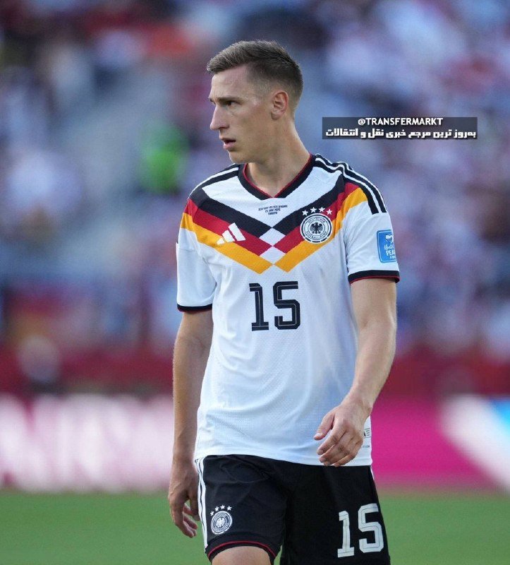 A soccer player wearing a white Germany national team jersey with the number 15 stands on a pitch during a match, with blurred stadium seating visible behind him.
