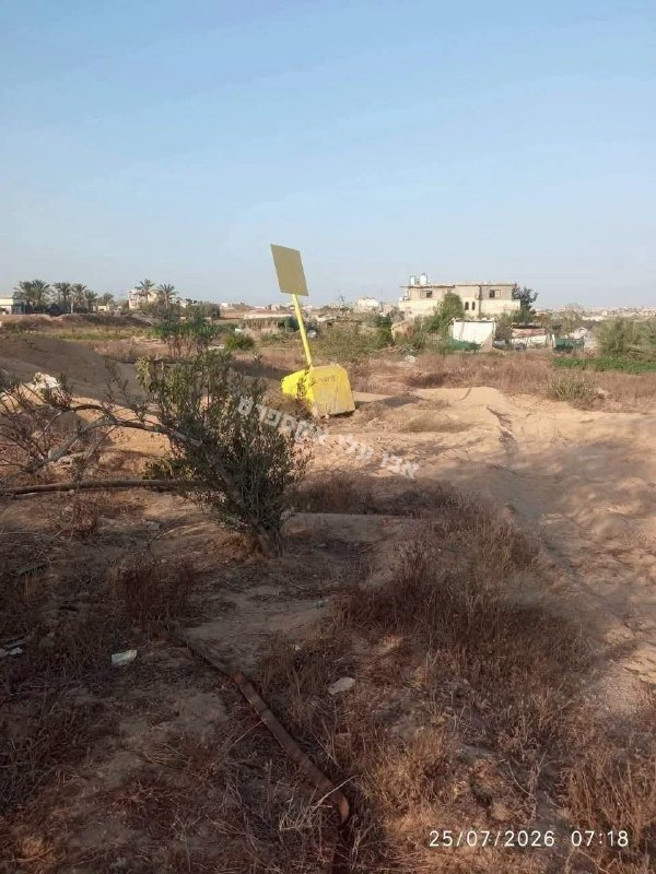 A yellow damaged signpost stands in a dry, sandy field scattered with sparse vegetation, with low buildings and palm trees visible in the distant background.