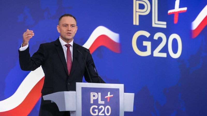 A man in a dark suit and red tie gestures while speaking at a podium labeled "PL+ G20" against a blue backdrop featuring the same logo.