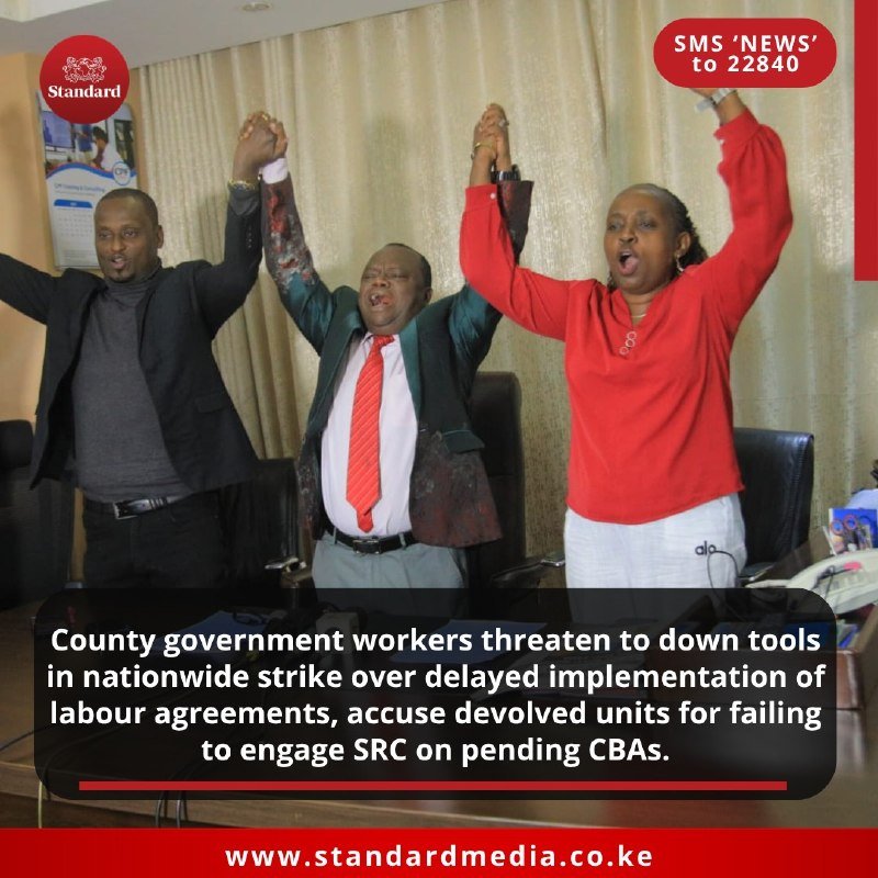 Three people stand side by side indoors with arms raised and joined hands, appearing to chant or protest.