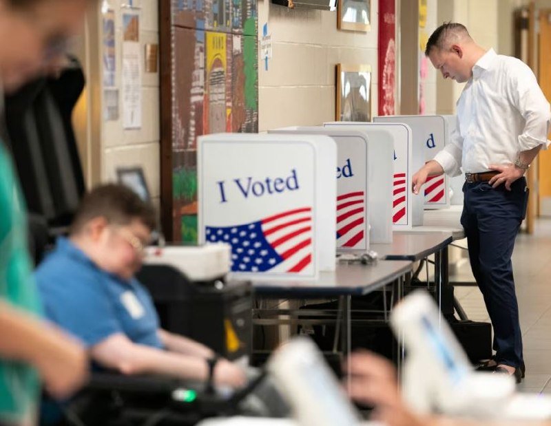 A man in a white shirt stands at an "I Voted" privacy booth inside a building with colorful artwork on the walls.