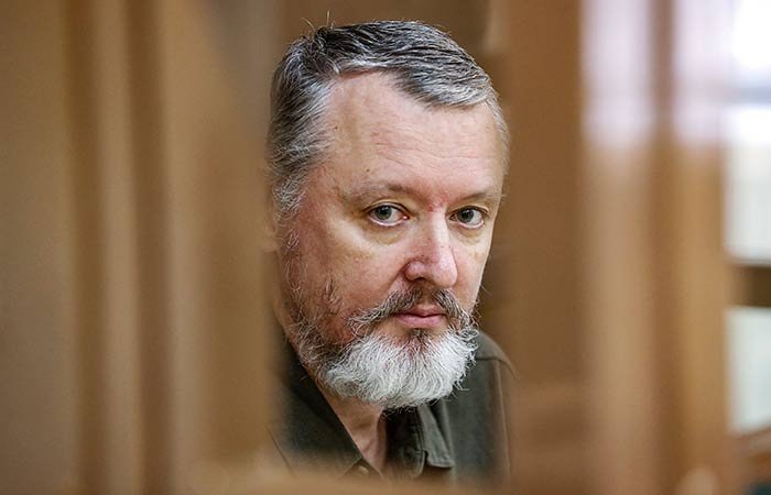 A bearded man with graying hair looks forward through an opening in a wooden structure, appearing to be in an indoor setting.