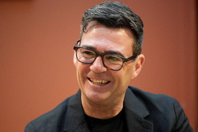 A smiling middle-aged man with dark hair and black-framed glasses wears a dark blazer over a black shirt against a solid terracotta-colored background.