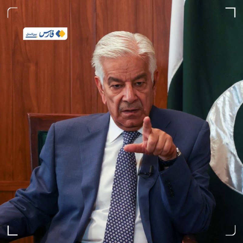 A white-haired man in a blue suit and patterned tie gestures with his hand while seated in front of a wooden wall and a green and white flag.