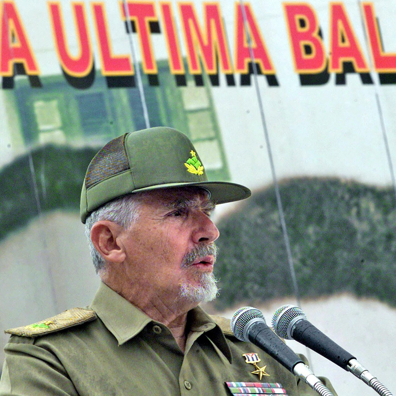 A gray-haired military officer in an olive-green uniform and cap speaks into two microphones before a large banner displaying a soldier and red lettering.