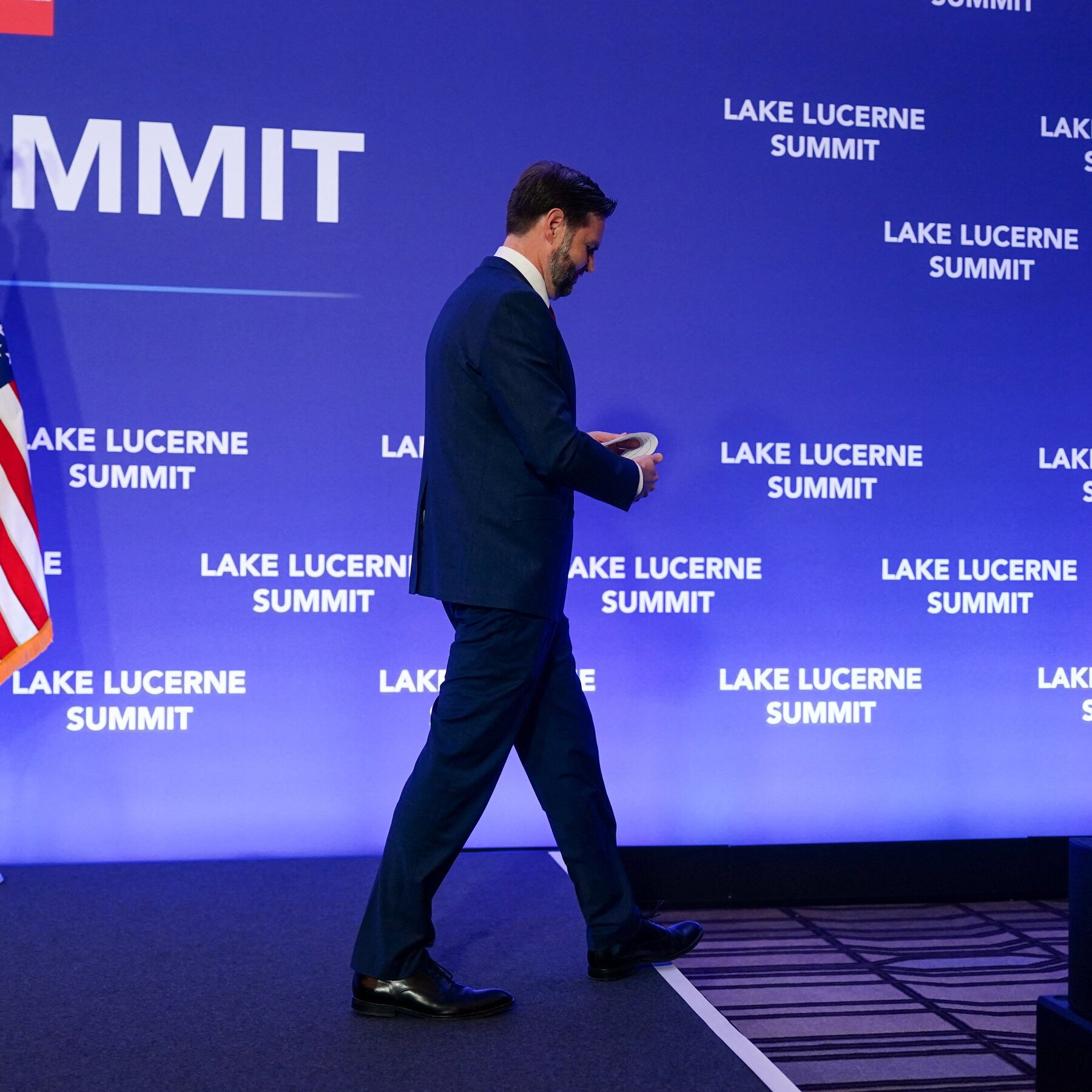 A man in a dark suit walks across a stage holding papers, with a blue backdrop displaying "LAKE LUCERNE SUMMIT" and "MMIT" logos.