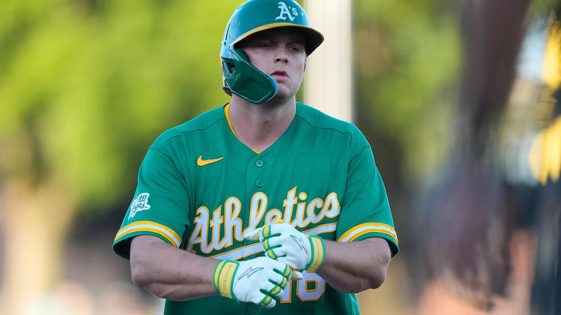 A baseball player in a green Oakland Athletics jersey and matching green helmet adjusts his batting gloves.