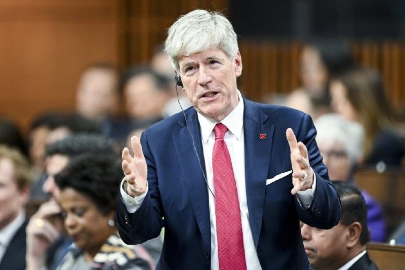 A gray-haired man in a navy suit and red tie gestures with both hands while wearing an earpiece in a crowded formal chamber.