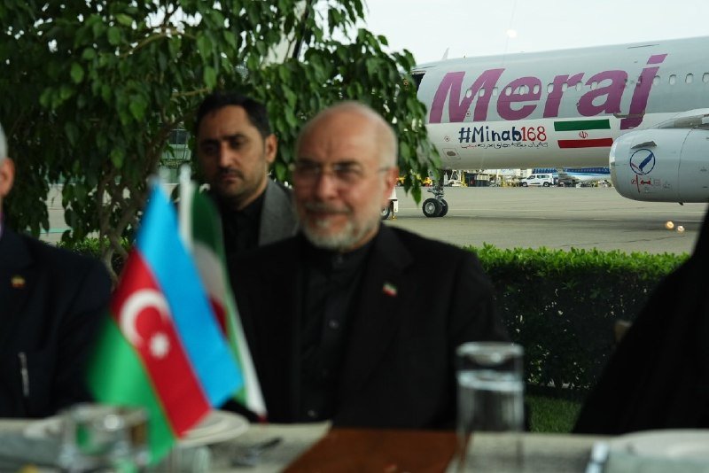 A man with a gray beard sits at an outdoor table near Azerbaijani and Iranian flags, with a Meraj airline aircraft visible in the background.