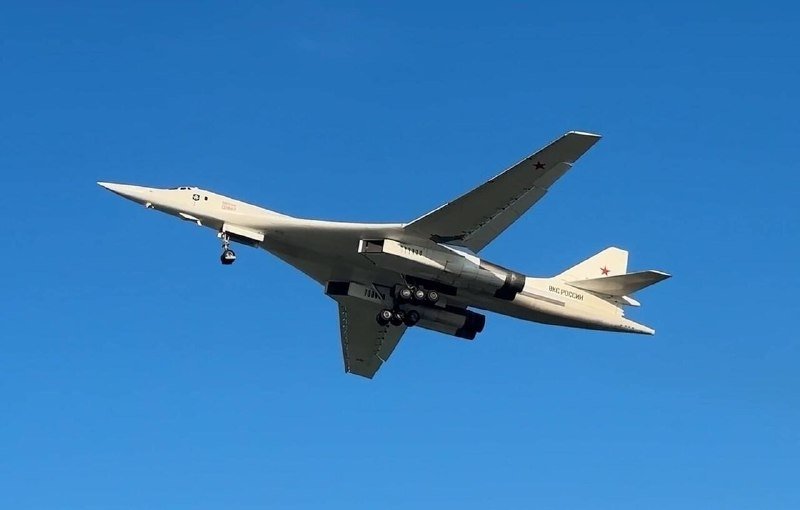A white military aircraft with swept wings, red star insignia, and "ВКС РОССИИ" markings flies against a clear blue sky.