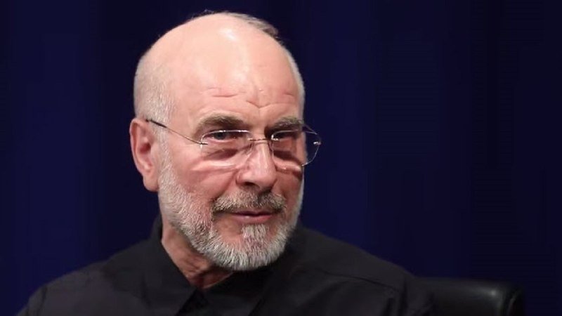 A bald, bearded older man wearing rimless glasses and a black shirt speaks against a dark blue background.