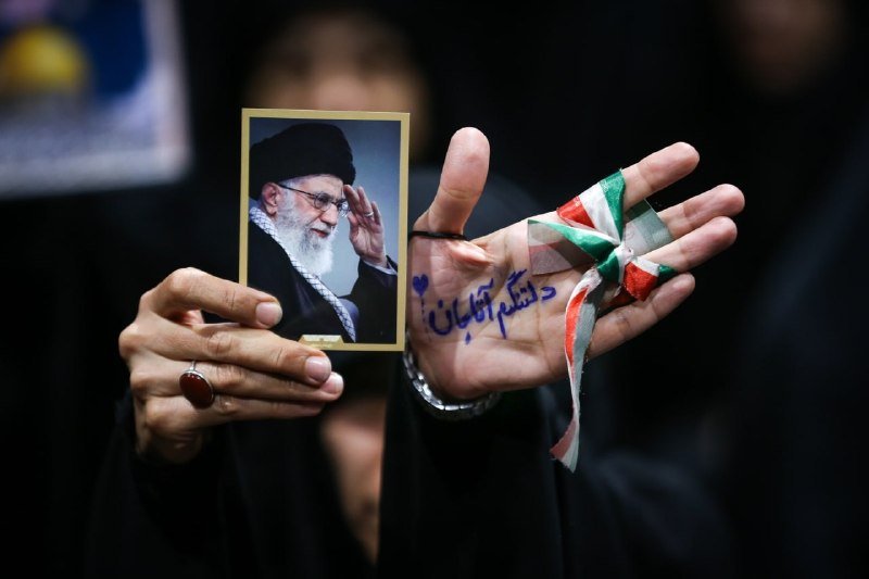 A person in dark clothing holds up a framed photograph of a bearded man wearing a black turban, alongside a hand with Arabic script and a green, white, and red ribbon tied to a finger.