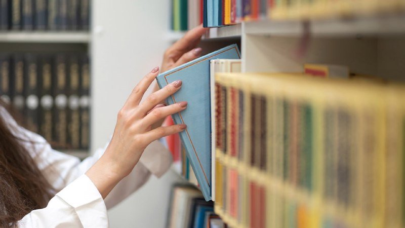 A person in a white shirt pulls a light blue book from a densely packed library shelf lined with rows of hardcover volumes.