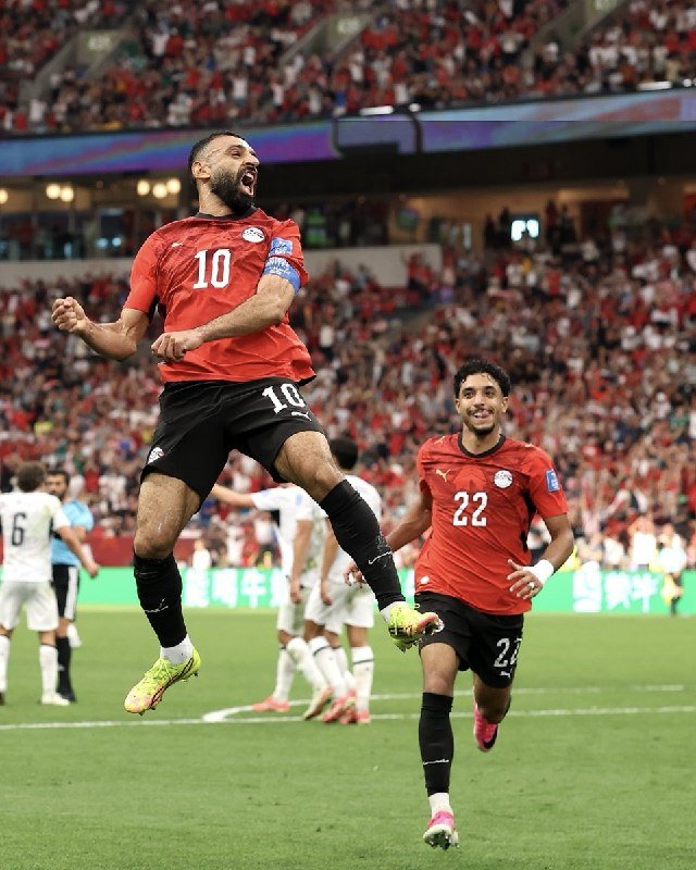 A bearded soccer player wearing a red jersey with the number 10 leaps into the air with clenched fists in celebration, while a teammate wearing number 22 runs toward him on a stadium field.